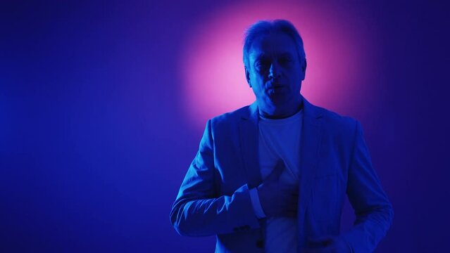 Heart Attack. Stressed Aged Man. Neon Light Portrait. Worried Male Holding Hand On Chest Suffering From Pain In Pink Blue Blinking Dark Background Halo Lens Flare.