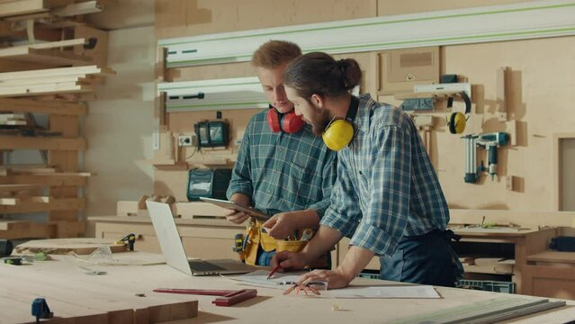 Small Business Owners of a Furniture Workshop Using Tablet, Notebook and Discussing the Design of a New Wooden Product. Handsome Carpenter and Young Male Apprentice Working in Studio.