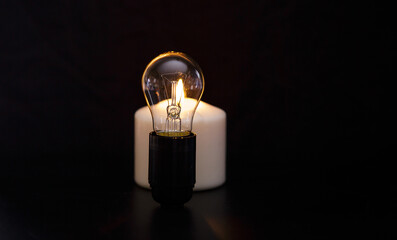 Burning candle near a switched off light bulb in dark home. Blackout, electricity off, load...
