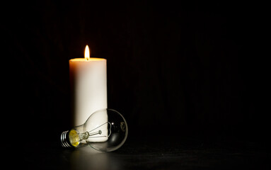 Burning candle near a switched off light bulb in dark home. Blackout, electricity off, load...