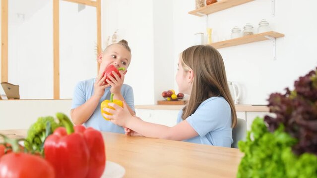 Little brother and sister in blue t-shirts are sitting in the kitchen and eating fresh vegetables. 