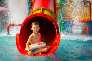 A cute little boy with an inflatable ring rides on a children's red water slide.