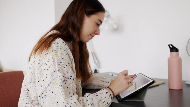 Side view of young woman student drawing on digital tablet doing homework, sitting by her dorm room desk