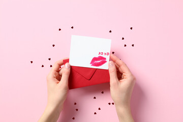 Valentine card mockup with lipstick kiss and red envelope in female hands over pastel pink background with confetti.