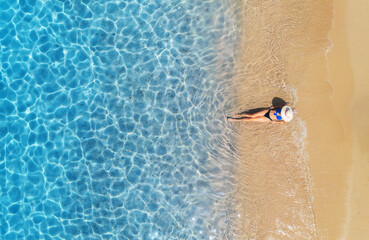 Aerial view of a lying woman in white hat on sandy beach and blue sea at sunny day in summer. Tropical landscape with girl, clear water, waves, sea coast. Top view. Vacation in Sardinia island, Italy	