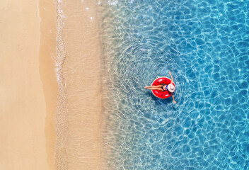 Aerial view of a woman in hat swimming with red swim ring in blue sea at sunrise in summer. Tropical landscape with girl, clear water, waves, sandy beach. Top view. Vacation. Sardinia island, Italy	