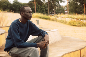 Obraz premium A dark-skinned young man sits on a wooden bench in the park and listens to music.