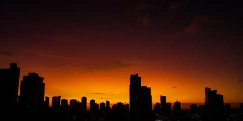 Urban sunset seen from Tirol, Natal, Rio Grande do Norte. Silhouettes of buildings formed by yellow-orange sunlight under a reddish sky.