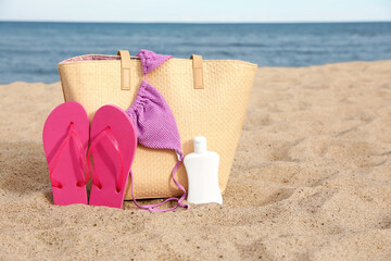 Summer bag with slippers, sunscreen and bikini top on sand near sea, space for text