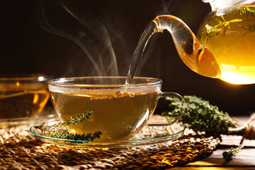 Pouring aromatic herbal tea into cup and thyme on wooden table - obrazy, fototapety, plakaty