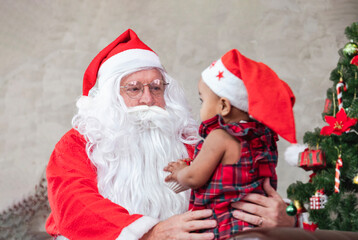 Santa Claus is lifting happy little toddler baby girl up and sitting on his lap with fully decorated christmas tree on the back for season celebration concept