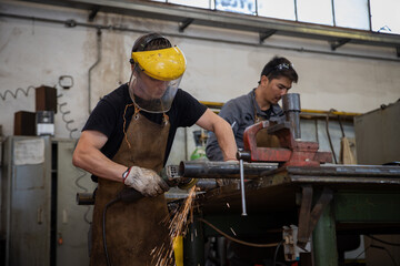 A worker polishes a piece of metal with the sander in the factory with his colleague working...