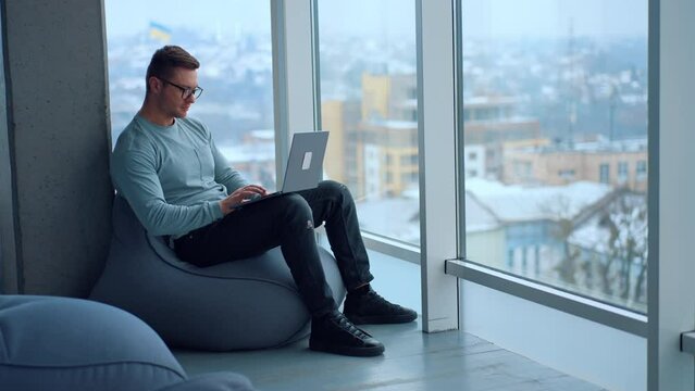 Male freelancer setting comfortably in bean bag chair in front of big panoramic windows. Man working on his laptop in a room with great view.