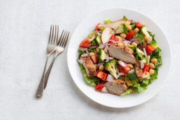 Healthy food salad bowl has chicken broccoli chickpeas tomato on white wood table.