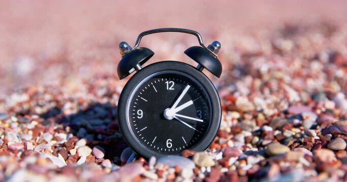 Alarm clock is on beach, small pebbles, second hand is ticking. Clock ticking fast time passing deadline. Distraction from bustle of meditation. Deadline inability to rest doing nothing on vacation 