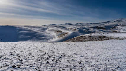 The dry grass in the endless snow-covered valley is covered with frost. In the distance, a winding trampled dirt road is visible between the hills. Blue sky. Altai expanses