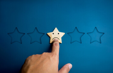 Sad emotion face on a wooden star pointing by hand on blue background. Negative reviews in customer...