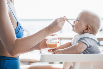 Mother spoon feeding her baby boy child in baby chair with fruit puree on a porch on summer...