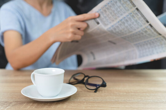 A cup of coffee,glasses on the wood table and woman read a newspaper.