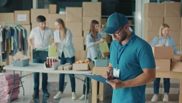 Male Volunteer Writing on Clipboard While Working in Food Bank Warehouse. Young Man Doing Inventory of Donated Goods in Volunteer Center While Group of Volunteers Sorting Humanitarian Aid