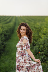 Naklejka na ściany i meble Happy young woman with brown curly hair, wearing a dress, posing outdoors in a garden