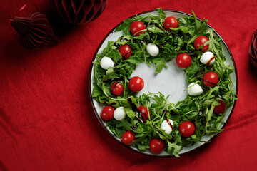 Christmas wreath of Salad with arugula, cherry tomatoes and mozzarella cheese on red cloth table with red balls. Christmas food background.