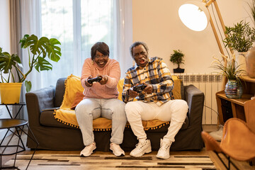 Obraz premium Funny senior African American couple sitting on the sofa playing video games with the game console controller