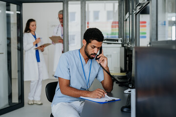 Young multiracial doctor sitting at reception and calling, his colleagues discussing in the background.