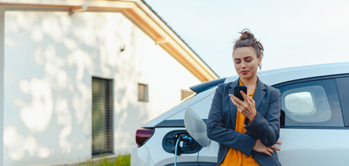 Young woman with smartphone waiting while her electric car charging in home charging station,...