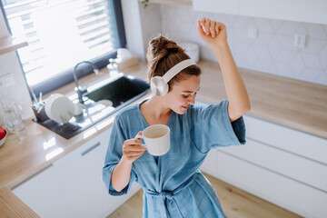 Young woman listening music and enjoying cup of coffee at morning, in her kitchen. - obrazy, fototapety, plakaty