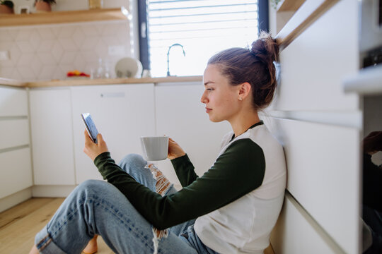 Young woman sitting with smartphone and cup of coffee in a kitchen.