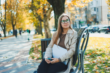 A cute fair-haired young woman is sitting in an autumn park on a bench and working on a laptop....