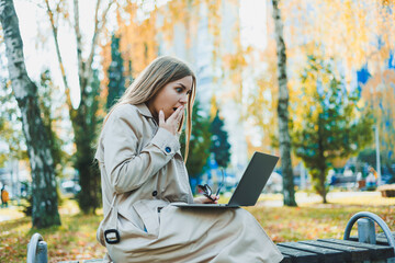 A cute fair-haired young woman is sitting in an autumn park on a bench and working on a laptop. Remote work