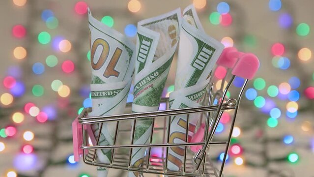 Paper banknotes dollars in a shopping cart on the background of Christmas lights. Place to copy. New Year multi-colored lights in defocus. Beautiful holiday card, video for intro, text, intro. UHD 4K.