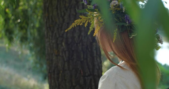 Beautiful blonde model posing  wearing  wreath of wild flowers on nature background. Close up portrait of young woman with long hair in rustic linen dress turning around looking to camera on sunny day