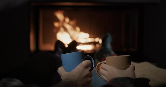 Hands of two young people with cups of tea on the background of a burning fireplace. Cozy and warm house concept