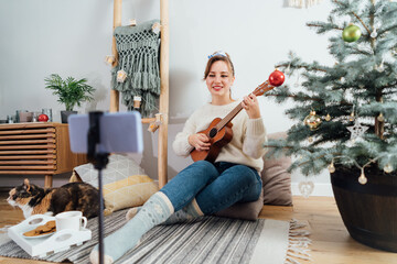 Young Woman playing ukulele guitar and recording Christmas music video for social networks on phone...