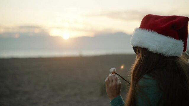 A girl in a Santa hat on the background of mountains and sunset eats fried marshmallows on the beach by the sea. New Year.
