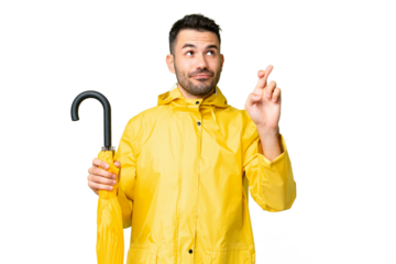 Young caucasian man with rainproof coat and umbrella over isolated background with fingers crossing and wishing the best