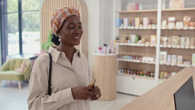 Happy young black woman paying by credit card for purchase on cashier counter, taking paperbag passed by shop assistant and leaving cosmetic supermarket