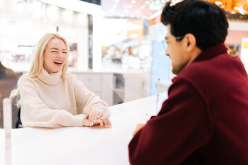 Back view from shoulder of unrecognizable young man chatting through glass partition with cheerful blonde woman standing in hall of shopping mall. Concept of lifestyle social distancing and work.