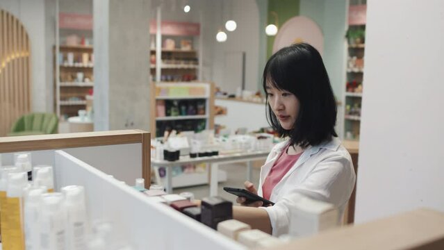 Young Asian consumer taking small bottles containing skincare products and scanning qr codes on them in cosmetic supermarket to learn online information about each item