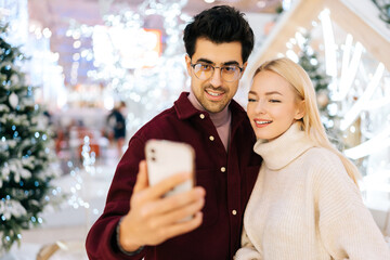 Close-up portrait of happy young couple taking selfie picture on mobile phone standing in hall of celebrate shopping mall in Christmas eve, on background of bright beautiful xmas decorations.