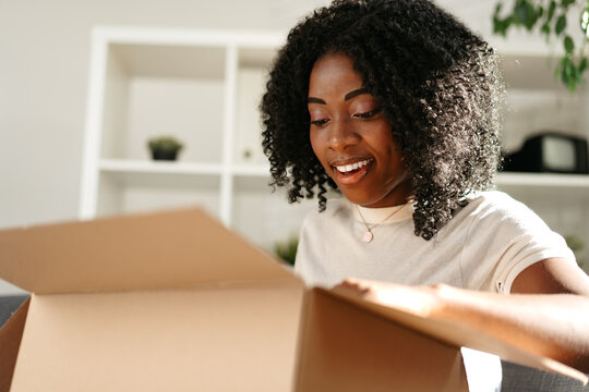 Young african woman sit on couch at home unpacking parcel cardboard box with online purchase