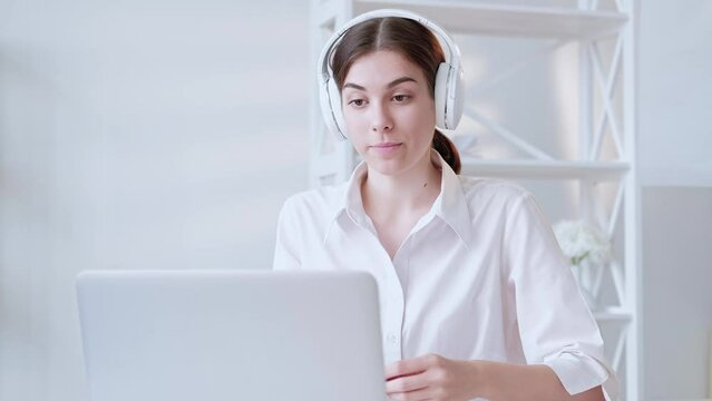 Video interview. Virtual meeting. Distance communication. Confident woman in headphones speaking online using laptop at light modern digital home office interior with free space.