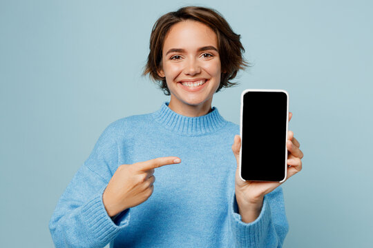 Young Smiling Woman Wears Knitted Sweater Hold In Hand Use Point Index Finger On Mobile Cell Phone With Blank Screen Workspace Area Isolated On Plain Pastel Light Blue Cyan Background Studio Portrait.