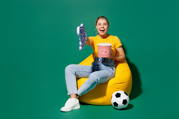 Fototapeta na wymiar Full body young woman fan wear t-shirt foam 1 fan glove finger up point camera on you cheer up football sport team sit in bag chair near soccer ball watch tv live stream isolated on green background.