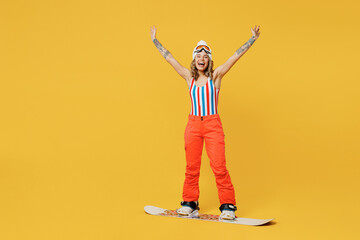 Snowboarder excited winner woman wear orange ski suit goggles mask hat swimsuit spend extreme weekend raise up hands isolated on plain yellow background studio. Winter sport hobby trip relax concept.