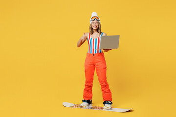 Snowboarder woman wear orange ski suit goggles mask hat swimsuit spend extreme weekend hold laptop pc computer show thumb up isolated on plain yellow background. Winter sport hobby trip relax concept.