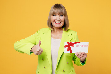 Elderly satisfied fun happy woman 50s year old wearing green jacket white t-shirt hold gift certificate coupon voucher card for store show thumb up isolated on plain yellow background studio portrait